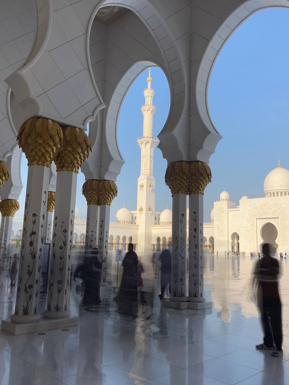 People walk through the Sheikh Zayed Grand Mosque corridors, ornate mosque with white and gold arches. A tall minaret rises against a clear blue sky, creating a serene atmosphere.