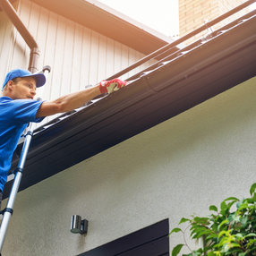 Man on ladder cleaning gutters, wearing a blue hat and gloves. Clear Choice.