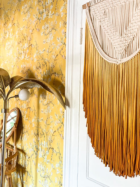 A close up photo of a yellow wallpapered wall in Room 6 with a macrame weaving and a gold palm tree