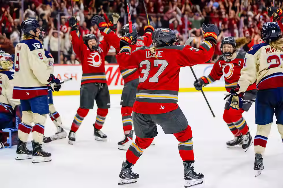 Four players from The Ottawa Charge celebrating the winning goal on the ice while players from the Montréal Victoire stand around.