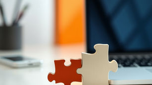 Two puzzle pieces, red and silver, connected on a desk with a laptop. Blurred orange object and phone in the background.