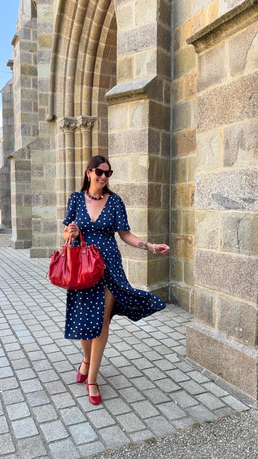 Jenny en robe bleu marine à pois, chaussures et sac rouges, devant un bâtiment en pierre