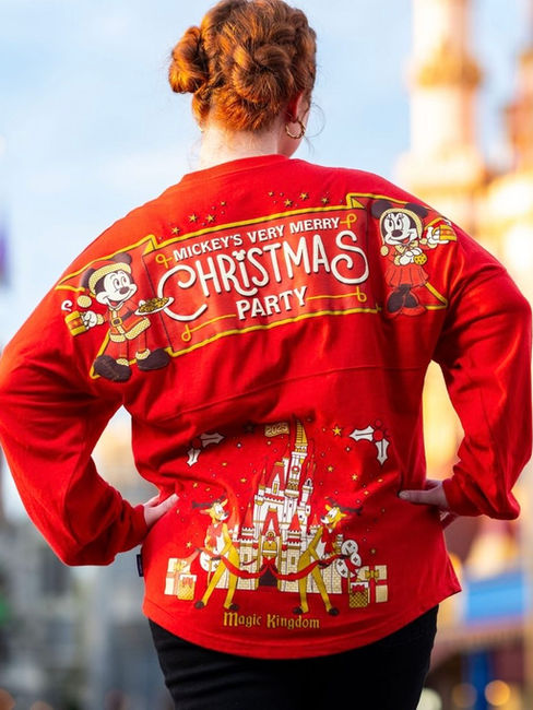 Person in red sweater with "Mickey's Very Merry Christmas Party" text, featuring Mickey and Minnie, stands in front of a blurred theme park.