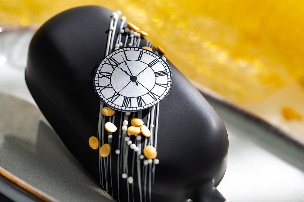 Black dessert with a clock face decoration, gold and white sprinkles on top, set against a bright yellow bokeh background.