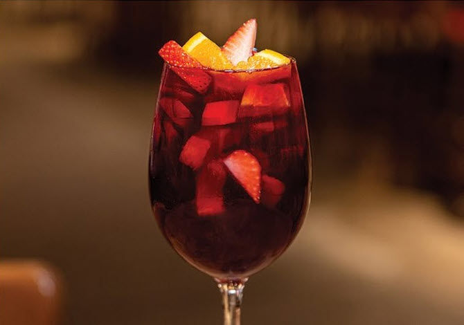 A glass of red sangria filled with ice, orange slices, and strawberries against a blurred, warm-toned background. Refreshing and vibrant.