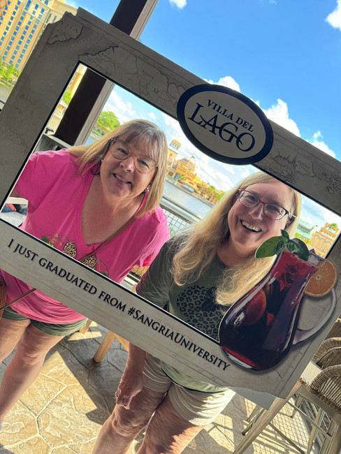 Two smiling women holding a "Villa del Lago" frame saying "I just graduated from #SangriaUniversity" with a lakeside backdrop.