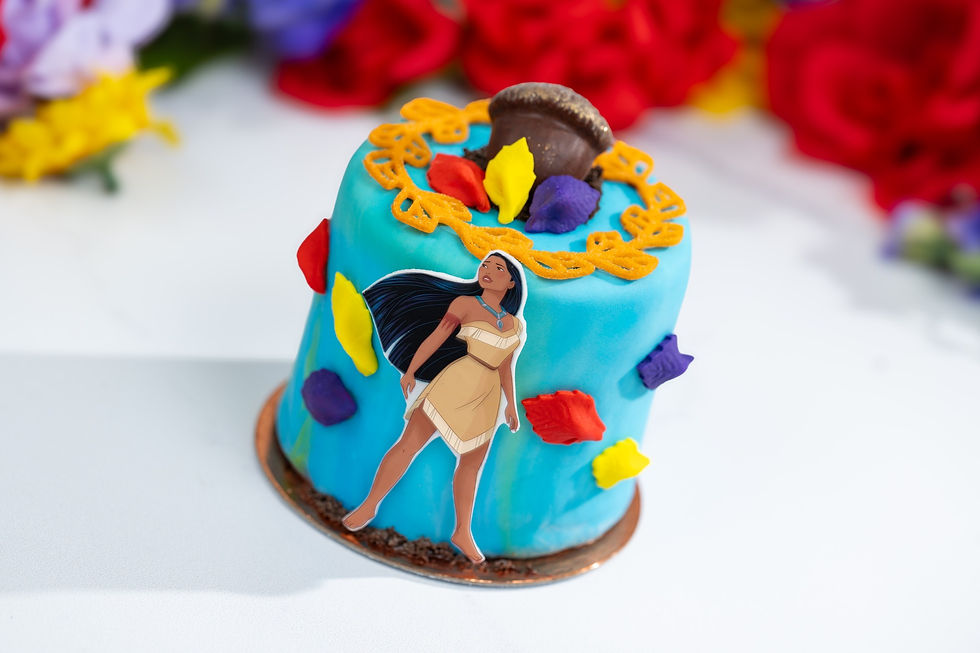 Blue cake with Pocahontas decoration, colorful leaves, and acorn on top. Red and yellow flowers blurred in the background.