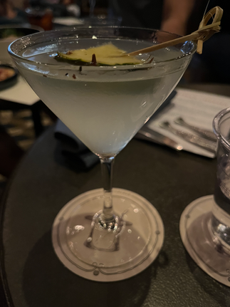 A martini glass with a light-colored cocktail, garnished with a pineapple slice. The setting is a dimly lit table, creating a relaxed mood.