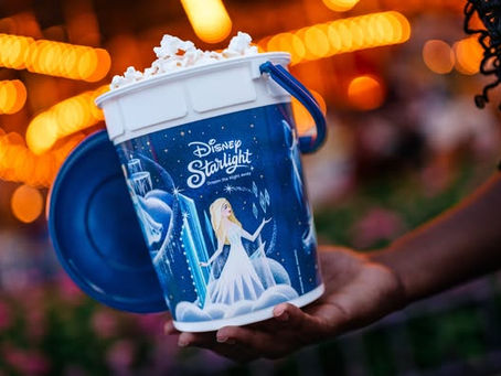 A person holds a blue and white Disney Starlight popcorn bucket with Elsa design. Warm lights glow in the blurred background, creating a festive mood.