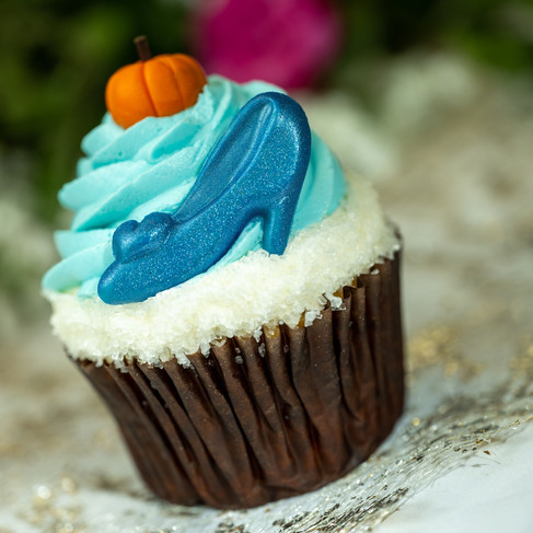 Cupcake with blue frosting, topped with an orange pumpkin and blue shoe. Background is blurred with pink and green hues. Playful mood.