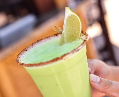A hand holds a green drink in a cup with a salt rim and lime wedge. Blurred warm background suggests an outdoor or casual setting.