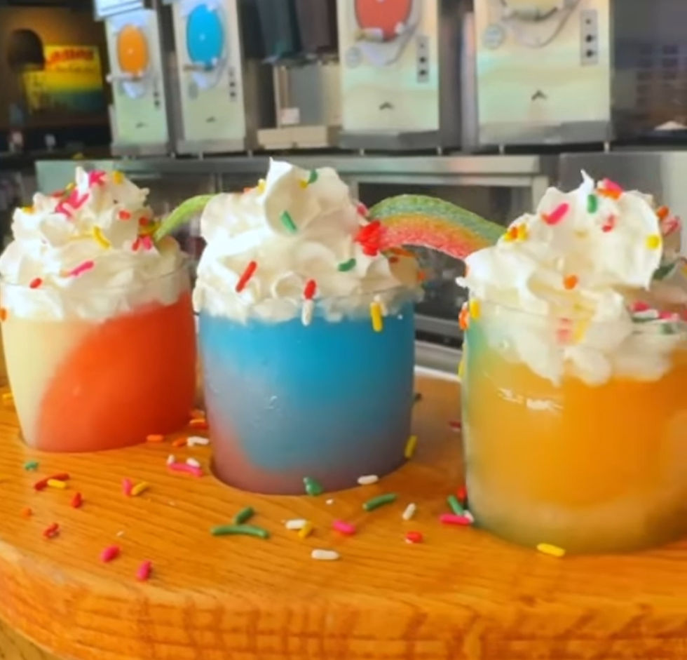 Three colorful slushies with whipped cream and sprinkles on a wooden counter. Machines in the background. Bright and playful mood. This is the Pride Flight at Splitsville at Disney Springs.