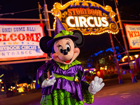 Character in a purple and green costume stands in front of "Storybook Circus" sign at night, with colorful lights and welcoming banners.