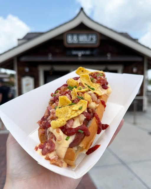 Close-up of a loaded hot dog topped with bacon, cheese, and chips. A hand holds it in front of a rustic building under a blue sky.