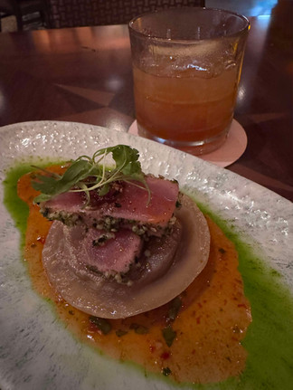 Seared tuna on a white dish with green and orange sauces, garnished with greens. A glass of brown drink on a coaster sits in the background.