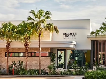 Modern building labeled "SUMMER HOUSE" surrounded by palm trees. Brick facade, large windows, sunny day, inviting atmosphere.