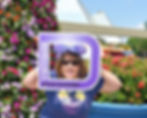 Woman smiling, holding a "Walt Disney World annual passholder" frame. She's wearing sunglasses and a purple bow, surrounded by vibrant flowers.