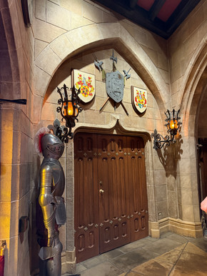 Armor-clad knight statue beside ornate wooden doors in a medieval-style room. Shields, coats of arms, and torch sconces adorn stone walls.