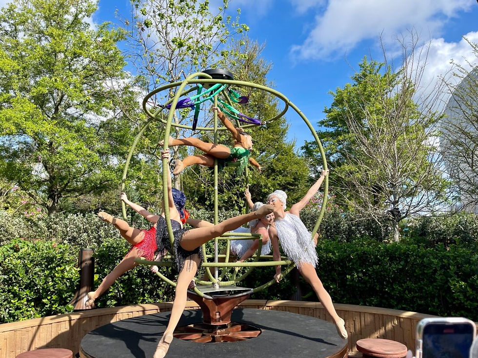 Acrobats in colorful costumes perform on a hoop structure in a garden setting. Green trees and a blue sky with clouds form the backdrop.