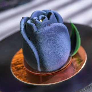 Blue rose-shaped dessert on a black plate with a golden base. Background has soft stripes, creating a sophisticated mood.