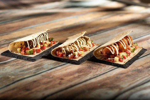 Three wraps filled with vibrant diced tomatoes, cucumbers, and meat, drizzled with sauce. Set on a rustic wooden table.