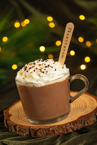 Glass mug of hot chocolate with whipped cream and sprinkles, garnished with a branded stirrer. Set on wood slice, festive bokeh lights.