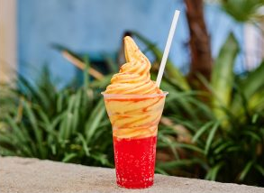 Swirled orange ice cream in a red cup with a straw, set against a lush green plant and blue wall background, creating a refreshing vibe.