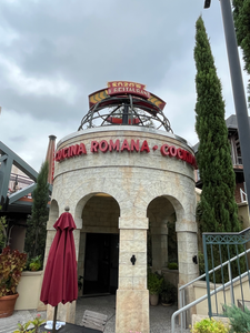 Stone rotunda with signs "Enzo's Restaurant" and "Cucina Romana Cocktails," under cloudy sky. Red umbrella and tall trees flank entrance.