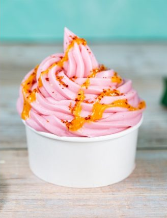 Pink soft serve ice cream swirled in a white cup, topped with orange sauce and sprinkles. Set against a light wooden background.
