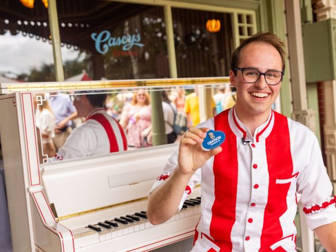 Smiling man in red-striped uniform holds a blue button at a white piano outside Casey's. Reflection and crowd visible in background.