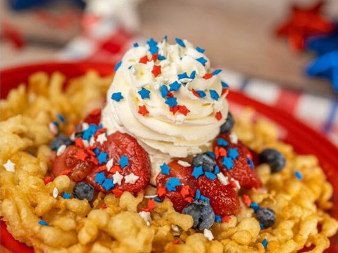 Funnel cake with whipped cream topped with blue and red star sprinkles, strawberries, and blueberries on a red plate, patriotic theme.