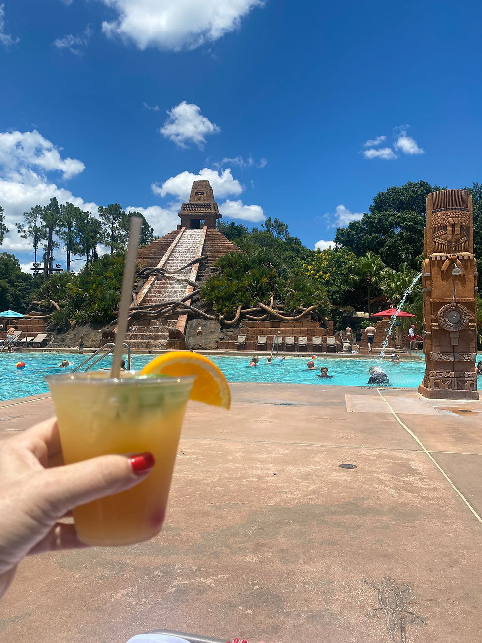 Hand holding a cocktail with an orange slice by a pool. Background features a pyramid with a waterfall, clear blue sky, and swimmer.