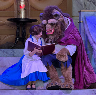 A woman in a blue dress reads a book with a beast in a red cloak. They sit on marble stairs, with a glowing rose in a glass case nearby.