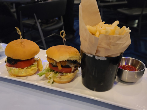Two mini burgers with lettuce and tomato, next to a cup of fries and ketchup on a white plate. Background shows empty chairs.