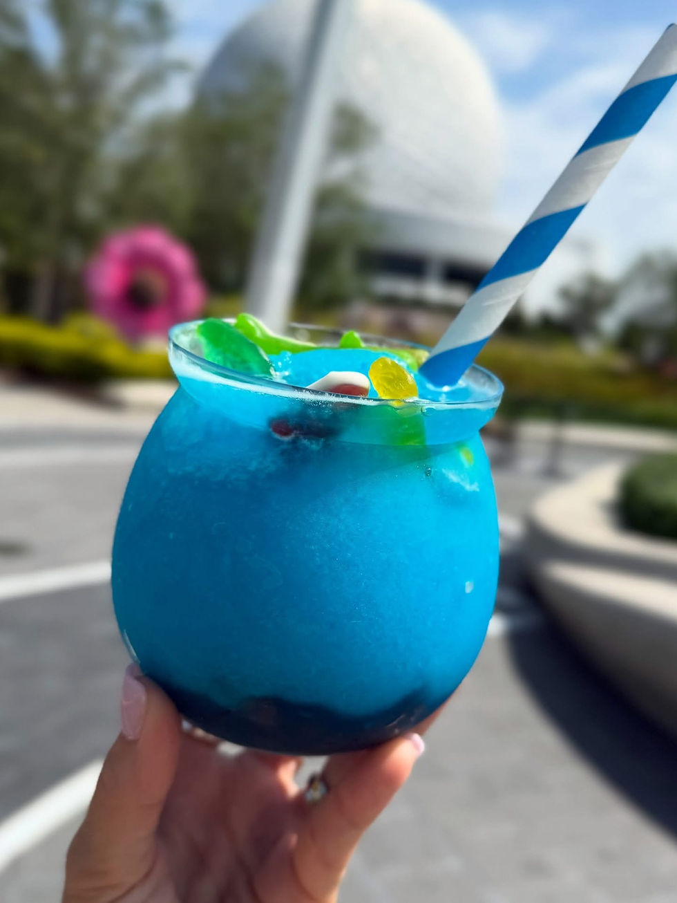 Hand holds icy blue drink with gummy candies and striped straw. Blurred background of a sphere and pink floral structure under blue sky.