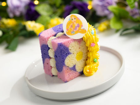Colorful floral cake slice with yellow icing on a plate, topped with an illustrated topper. Blurred floral background. Whimsical vibe.
