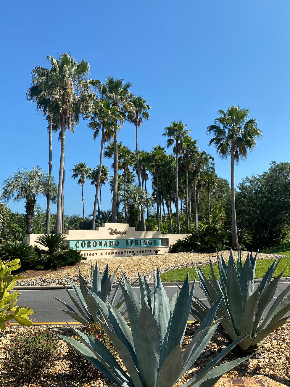 Tall palm trees and large agave plants surround a sign reading "Disney’s Coronado Springs Resort & Convention Center" under a clear blue sky.