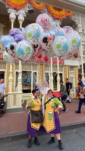 Two people in colorful uniforms hold Disney-themed balloons on a lively street. Bright banners and window displays form the festive backdrop.