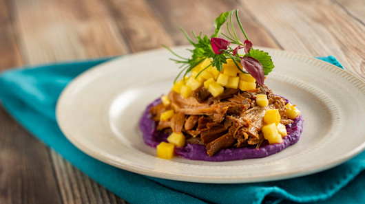 Pulled pork on purple puree, topped with mango cubes and greens, on a beige plate with a turquoise napkin, on a wooden table.