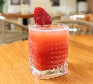Red drink with strawberry garnish in a textured glass on a wooden table. Blurred cozy café setting in the background.