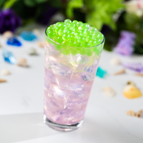 A pastel pink drink with ice and green boba in a glass. Background features colorful shells and greenery, creating a fresh vibe.