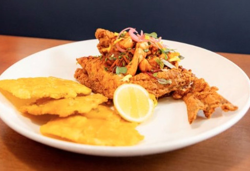 Fried fish with vegetables and a lemon wedge on a white plate, served with three golden, crispy tostones. Brown table background.