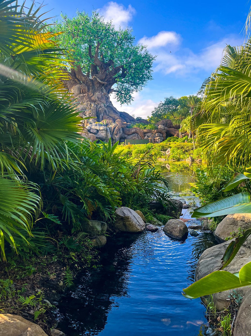 A lush scene with a large artificial tree, surrounded by greenery. A clear stream flows in the foreground under a vibrant blue sky.