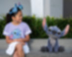 Girl in a purple Stitch shirt smiles at a plush Stitch toy on a bench, surrounded by green plants. Playful, joyful atmosphere.