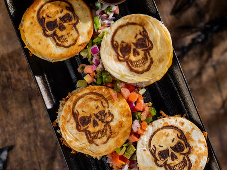 Four quesadillas with skull designs, surrounded by colorful diced vegetables on a black tray. A small bowl of red sauce is beside them.