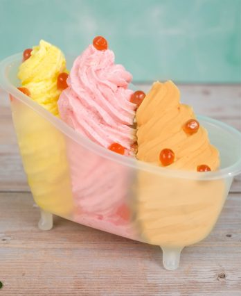 Three swirled ice cream scoops in yellow, pink, and orange with red cherries in a tiny bathtub on a wooden surface, teal background.
