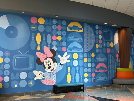 Colorful mural with a cartoon mouse character waving, surrounded by retro TVs, cars, and patterns. Bright, playful mood in a lobby setting.