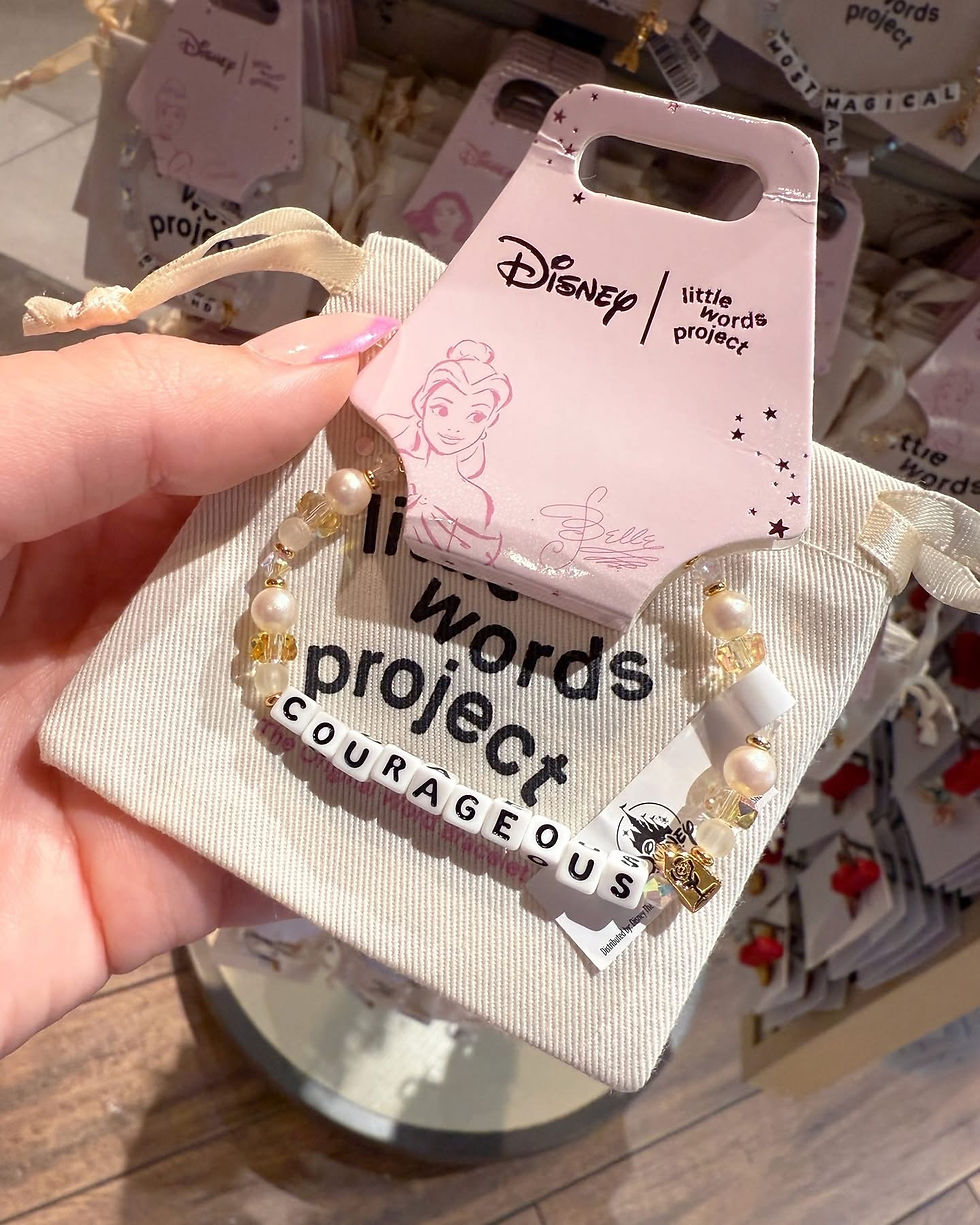 Hand holds a "Disney | little words project" bracelet reading "COURAGEOUS" with pearls, over a beige pouch. Belle illustration on tag.