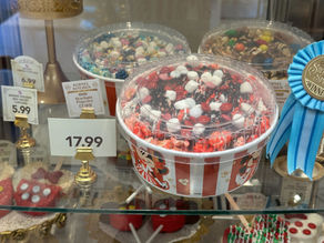 Red and white striped popcorn tubs with price tags and a blue ribbon in a display case. Colorful toppings visible on some containers.