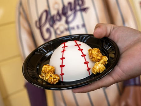 Hand holds a black plate with a baseball-themed dessert and caramel popcorn. A striped shirt with "Casey's Corner" is visible in the background.